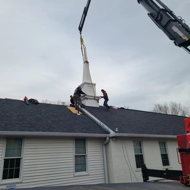 A boom crane lifting a new steeple onto the roof of Ardsley Bible Chapel while the Aronson crew guides it into place